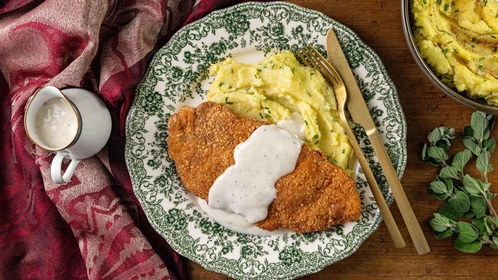 Breaded fried steak with white gravy, mashed potatoes with herbs, a fork, knife, and small gravy pitcher on a plate.