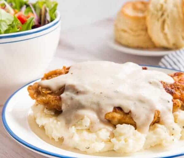 Chicken fried steak with white gravy over mashed potatoes, a biscuit on the side, and a bowl of salad in the background.