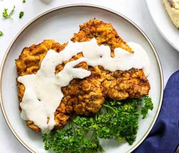Chicken fried steak with white gravy and parsley on a plate, accompanied by mashed potatoes, fork, and knife.
