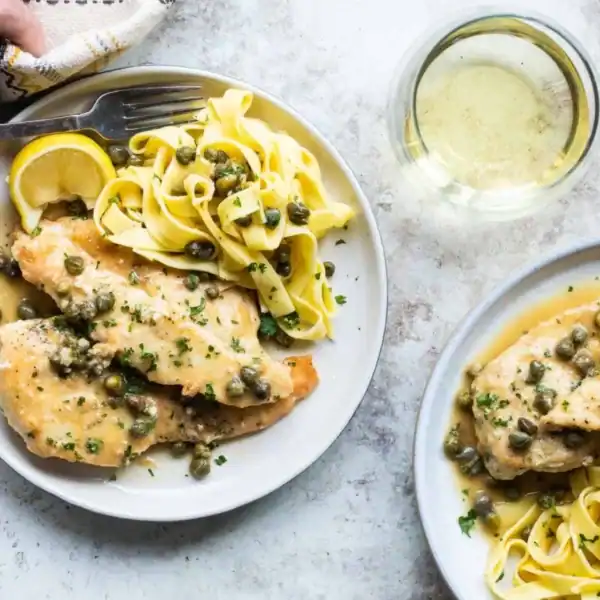 Two plates of chicken piccata with capers, lemon wedges, pasta, and a glass of white wine on a light-colored surface.