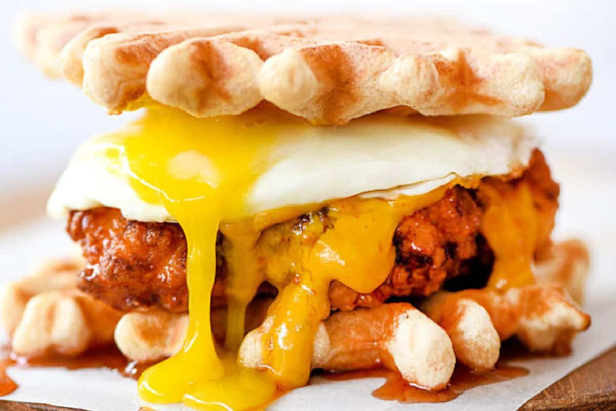 A sandwich made with waffles as the buns, featuring a fried chicken patty and a sunny-side-up egg with the yolk dripping. The golden yolk runs down the chicken and onto the waffles, creating a visually appetizing and indulgent dish.