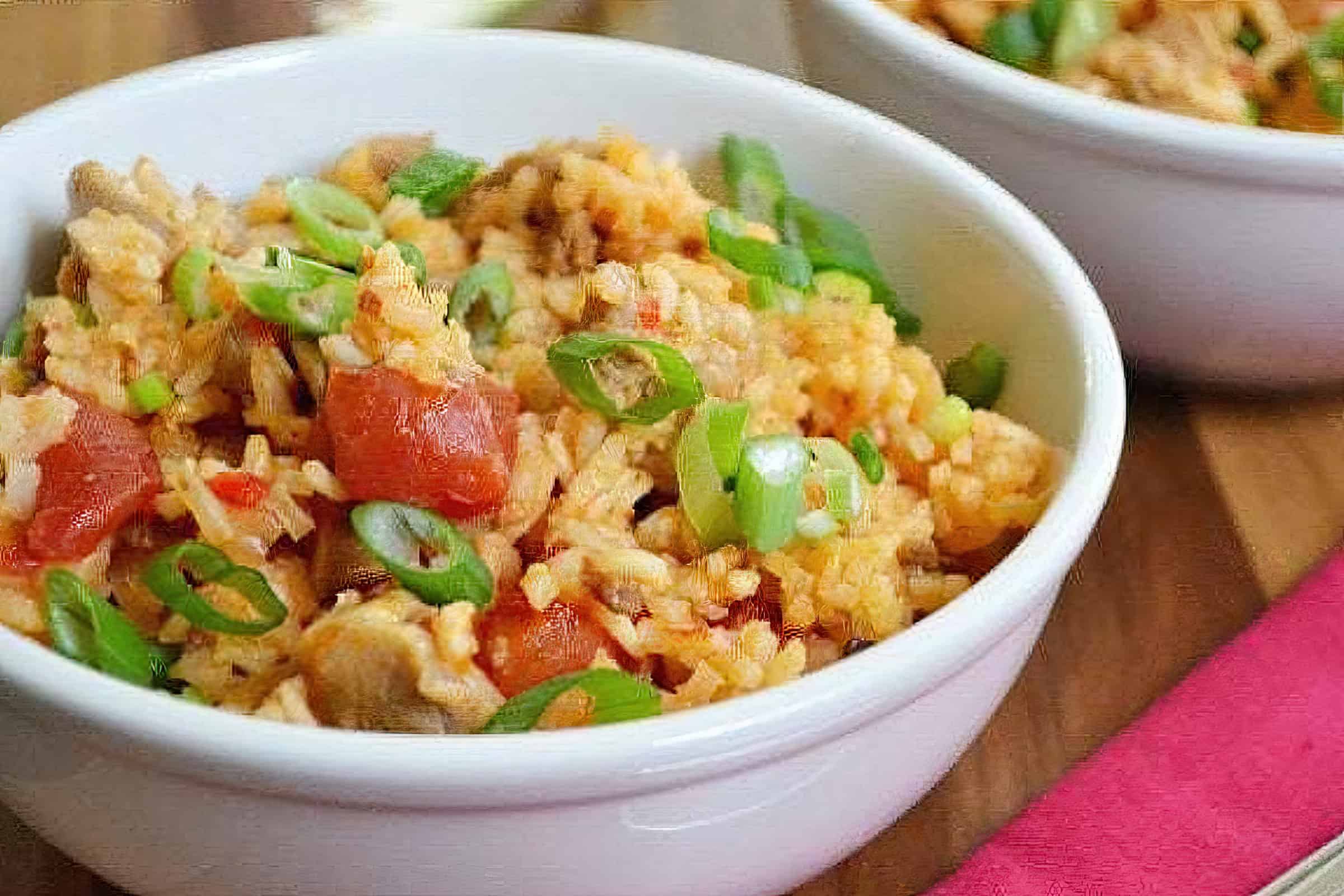 Two white bowls filled with flavorful rice mixed with chunks of tomatoes, diced green onions, and pieces of meat sit on a wooden table. The dish is garnished with fresh green onion slices, adding a burst of color to the hearty meal.