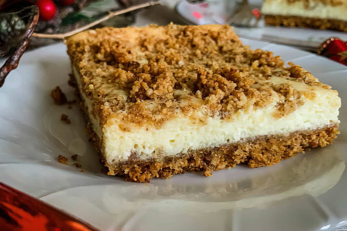 Close-up of a slice of crumb cheesecake on a white plate. The cheesecake has a creamy yellow layer topped with a golden brown crumb topping and a brown crust at the base. The background includes festive holiday decorations.