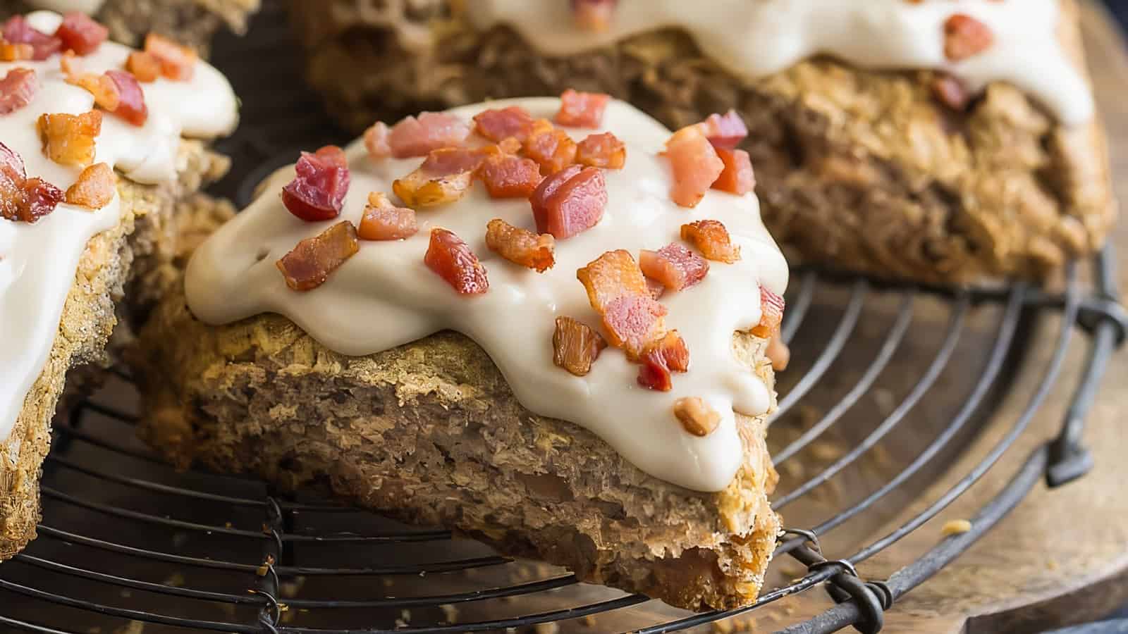 A close-up of a slice of dessert featuring a crumbly base topped with white icing and small bacon bits. The slice is placed on a black wire cooling rack, with other similar slices visible in the background.