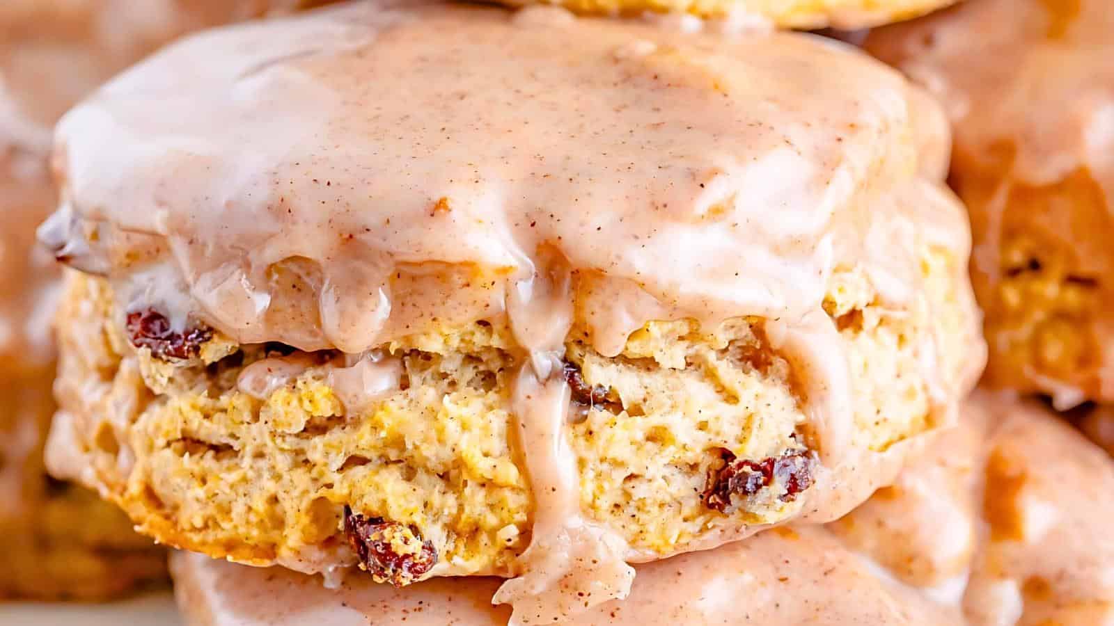 A close-up of a glazed biscuit with small pieces of dried fruit. The biscuit appears fluffy with a golden-brown exterior, and a generous coating of a light pink icing with specks of cinnamon drips down its sides. Other similarly glazed biscuits are in the background.
