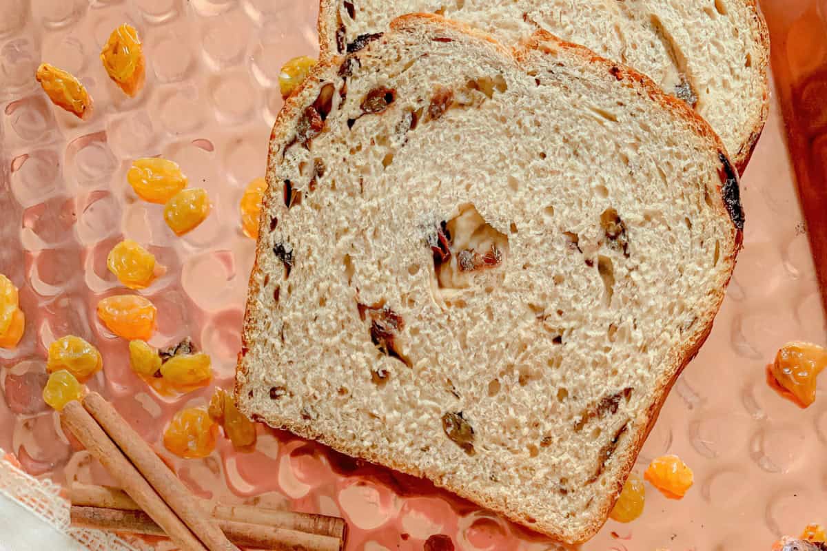 Two slices of raisin cinnamon bread lie on a textured copper background, surrounded by scattered golden raisins and cinnamon sticks. The bread has a light, fluffy look with visible raisins and swirls of cinnamon throughout.