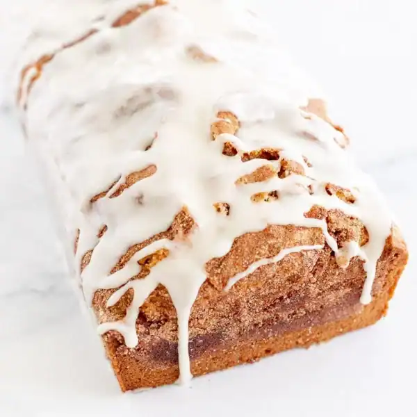A loaf of cinnamon bread with white icing sits on a white surface.