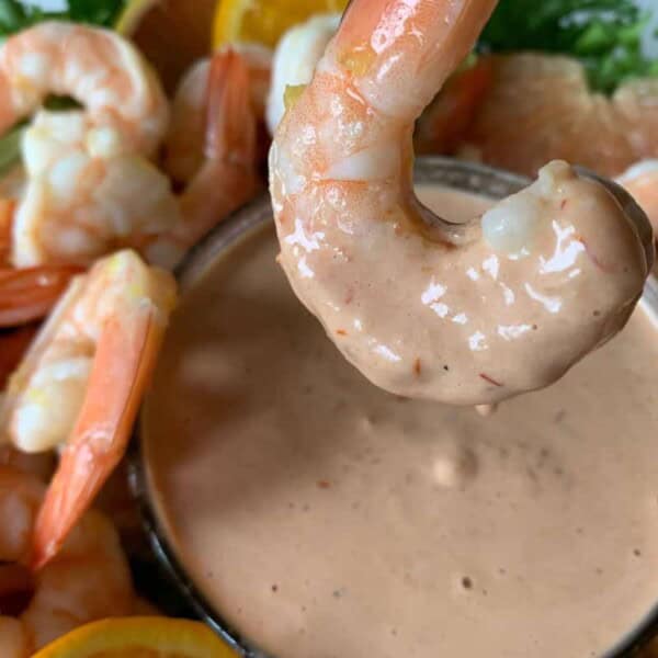 A shrimp dipped in a creamy sauce, with additional shrimp and lemon wedges arranged around the dip. Served with