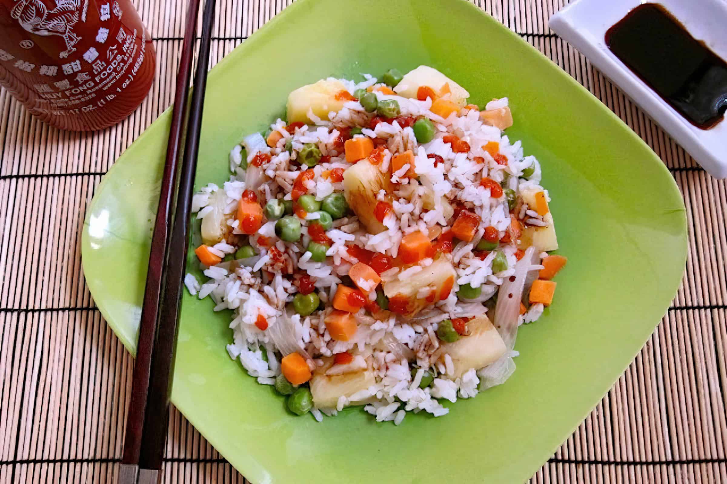 A green plate filled with colorful fried rice, including diced carrots, peas, corn, pineapple chunks, and small meat pieces. A pair of chopsticks rest on the plate's edge. A bottle of hot sauce and a small dish of soy sauce are placed nearby.