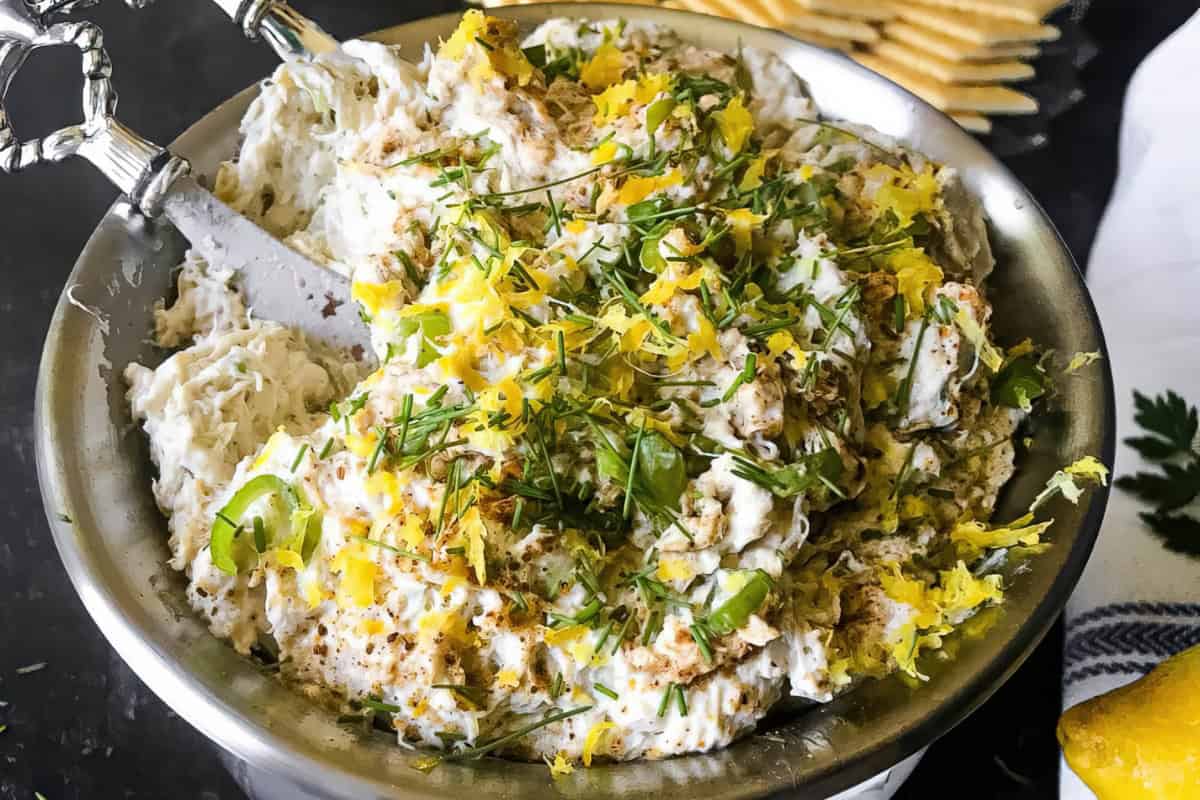 A stainless steel bowl filled with a creamy dip, garnished with chopped green herbs and lemon zest. A silver spreader knife with an ornate handle rests in the bowl. Crackers are partially visible in the background on the right side.