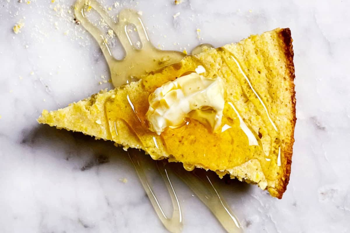 A slice of cornbread topped with a dollop of butter and drizzled with honey, sitting on a white marble surface. The honey is visibly flowing over the cornbread edges, creating a glossy, appetizing effect. Crumbs are scattered around the slice.
