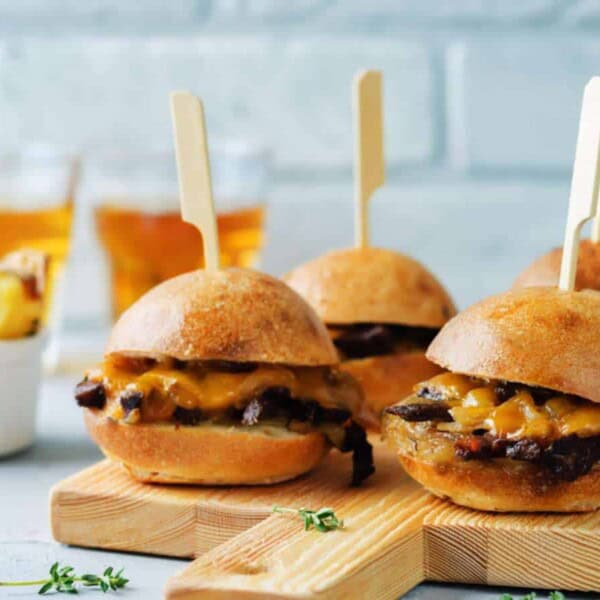Four cheeseburger sliders with wooden picks on a wooden board, accompanied by a bowl of potato wedges and two glasses of beverage in the background.