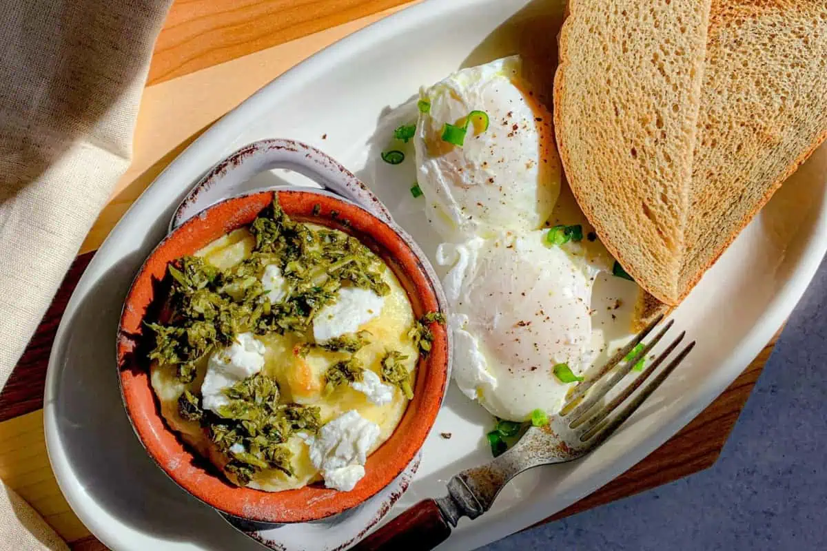 A white oval plate holds two slices of toast, two poached eggs garnished with green herbs, and a small brown dish filled with a mixture of greens and crumbled white cheese. A fork rests on the plate. The meal is set on a wooden surface with a beige napkin nearby.