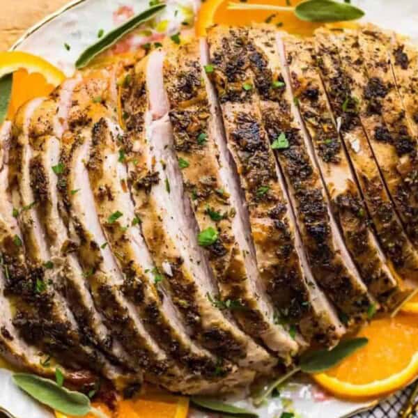 Sliced herb-crusted roast pork garnished with fresh orange slices and herbs on a white platter.