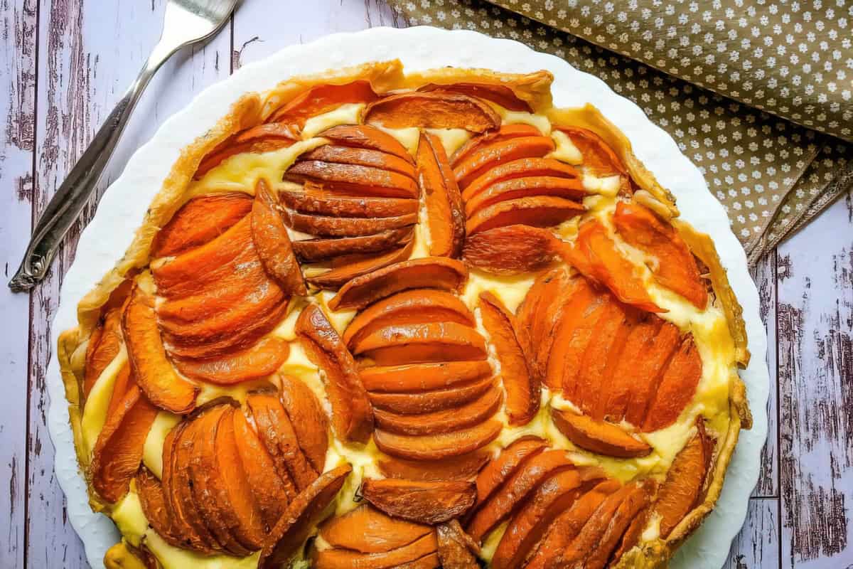 A baked sweet potato and cream tart with a flaky crust, neatly arranged on a white plate. The tart is decorated with thinly sliced, golden-brown sweet potatoes in a circular pattern. A fork and brown polka dot napkin are placed beside the plate on a wooden surface.