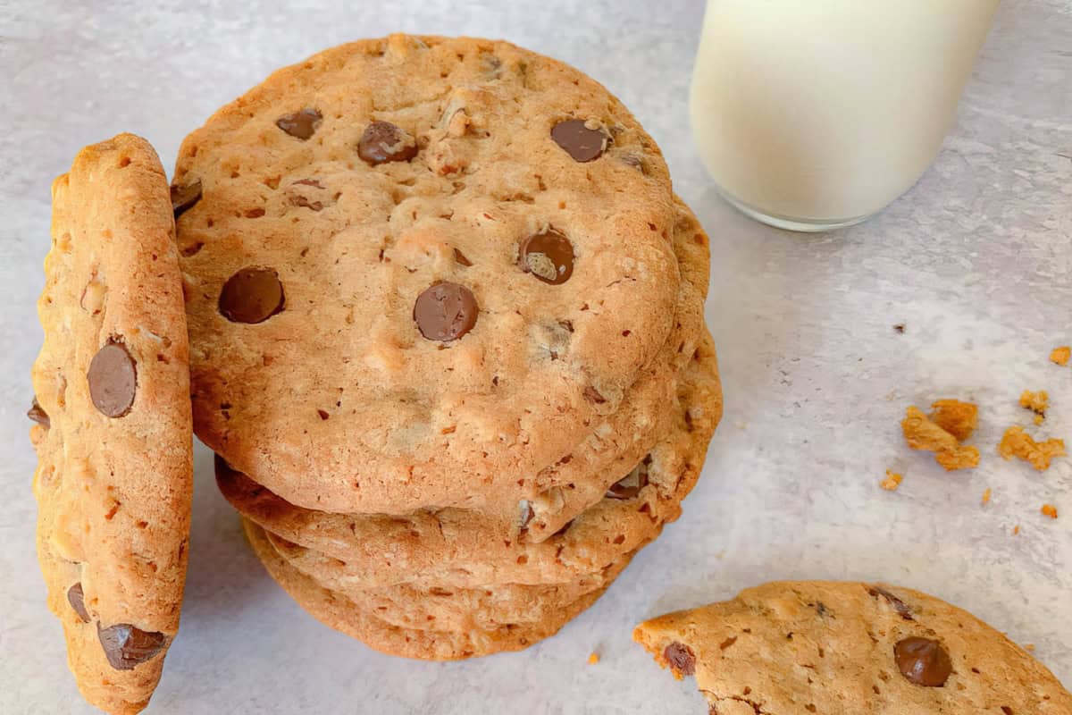 Chocolate chip cookies are stacked on a light surface beside a glass of milk. One cookie is leaning against the stack, and a broken cookie with crumbs is in the foreground.