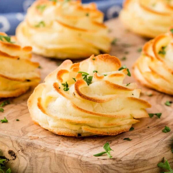 Four baked duchess potatoes with chopped herbs sit on a wooden board, accompanied by fresh greens.