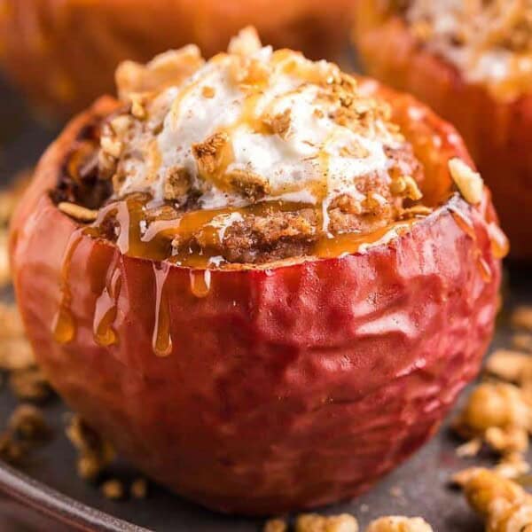 A baked apple stuffed with crumble, topped with whipped cream and caramel sauce, on a plate with scattered granola.