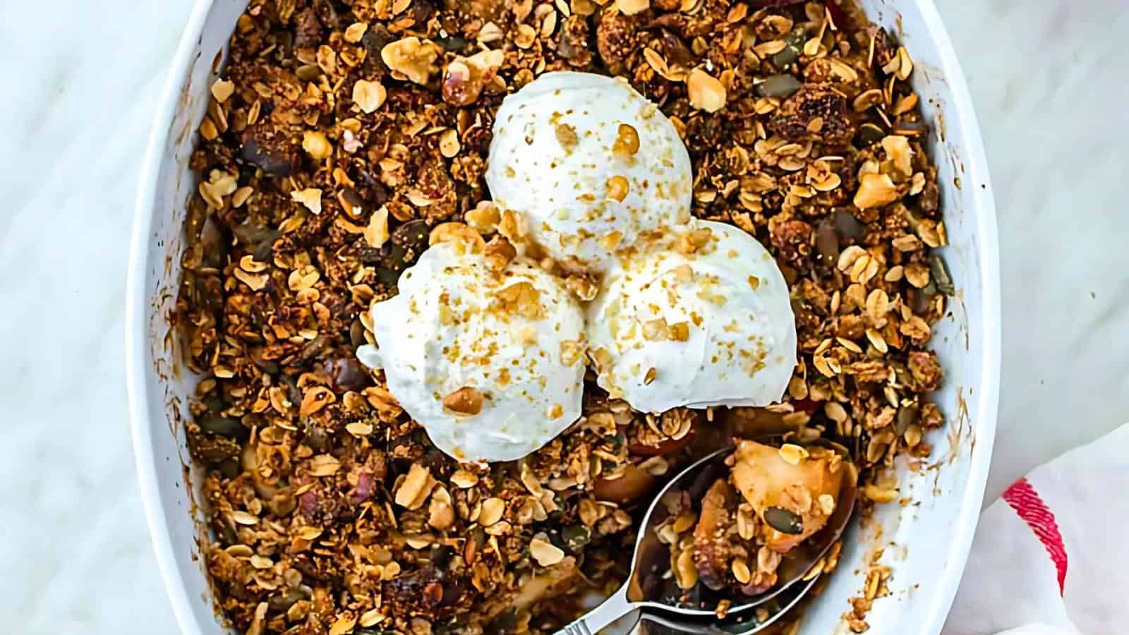 A baked apple crumble is shown in a white dish, topped with three scoops of vanilla ice cream. The crumble features a crispy golden brown oat and nut topping, with a serving spoon resting inside the dish on the right side.