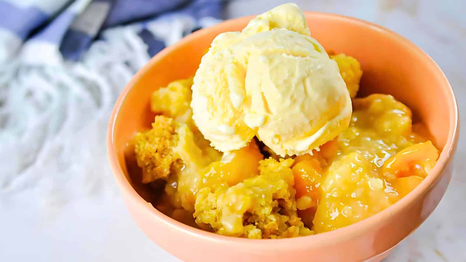 A peach cobbler served in a peach-colored bowl is topped with a creamy scoop of vanilla ice cream. The cobbler features chunks of peaches and a golden crust. A blurred white and blue cloth is visible in the background.