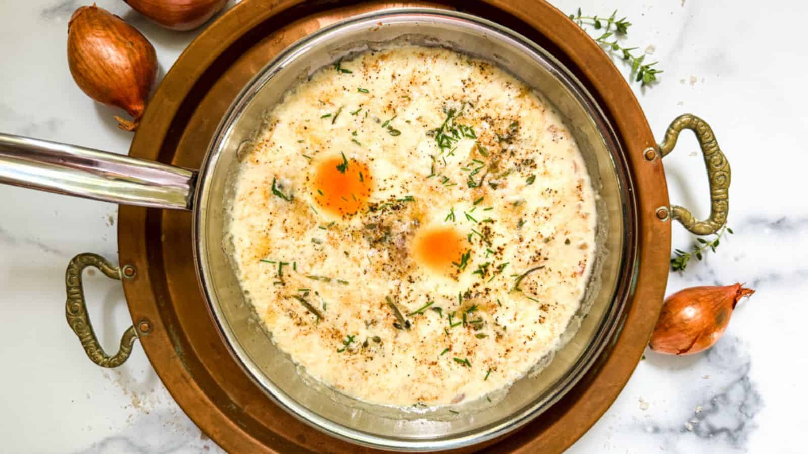 A skillet containing a creamy grits dish topped with two soft-cooked eggs, garnished with herbs and black pepper. The skillet is placed on a decorative brass tray, with unpeeled onions and sprigs of thyme on the side on a marble countertop.