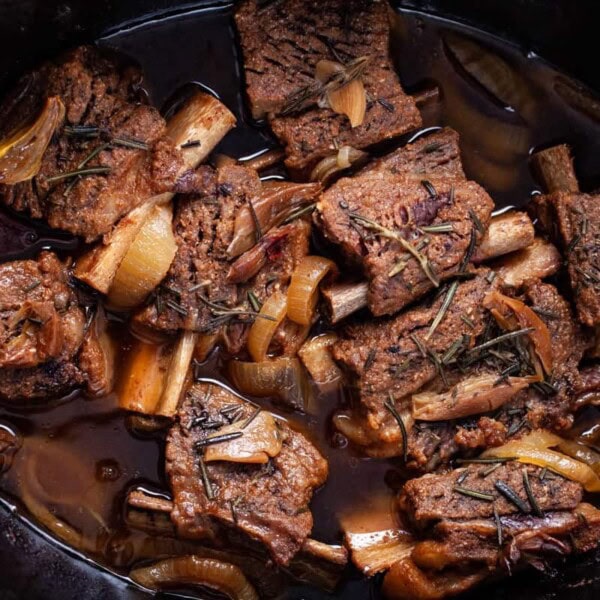 A slow cooker filled with braised short ribs, onions, and herbs in a dark, rich sauce.