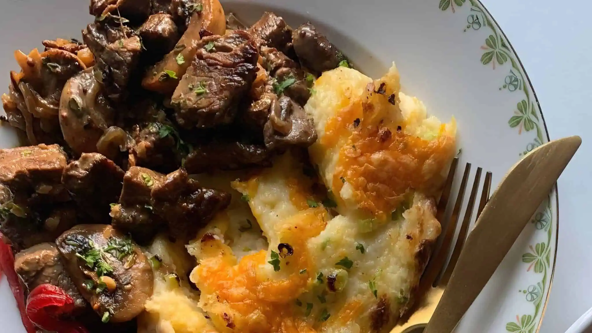 A plate of creamy mashed potatoes topped with melted cheese alongside a serving of savory beef and mushroom stew. The dish is garnished with chopped herbs and accompanied by a fork, all served on a white plate with a green floral pattern on the rim.