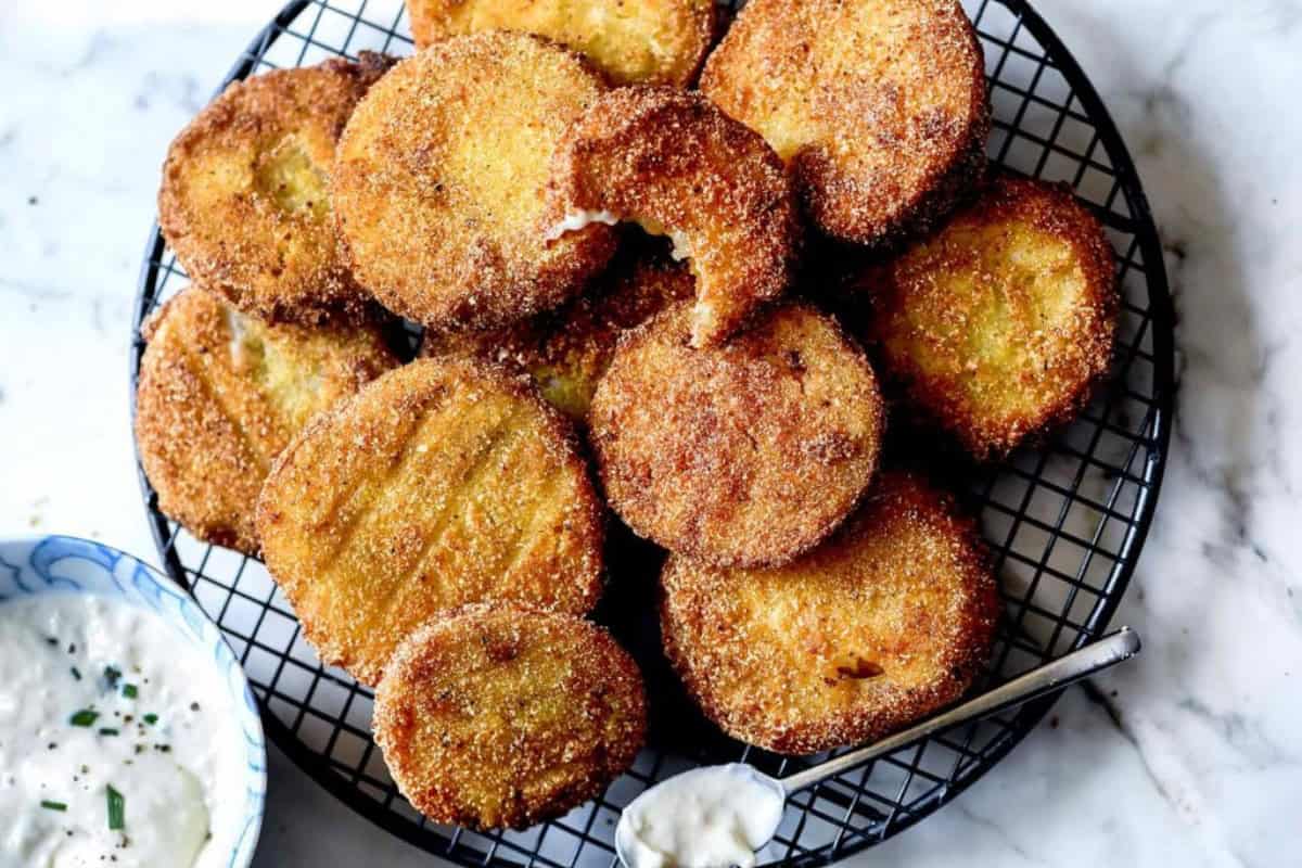 A plate of golden-brown fried green tomatoes arranged on a black wire cooling rack. One tomato has a bite taken out of it, showcasing the crispy texture. A small bowl of creamy dipping sauce with green herbs is placed beside the plate.