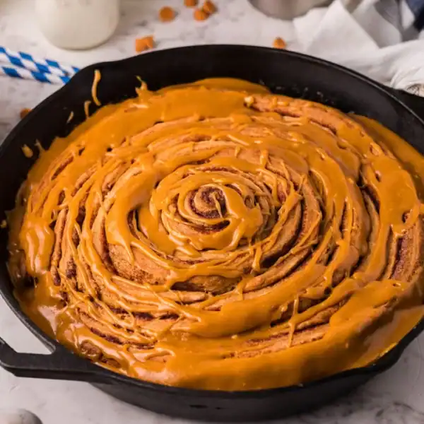 A large cinnamon roll with caramel icing in a cast iron skillet on marble, next to a bowl of caramel sauce and butterscotch chips.