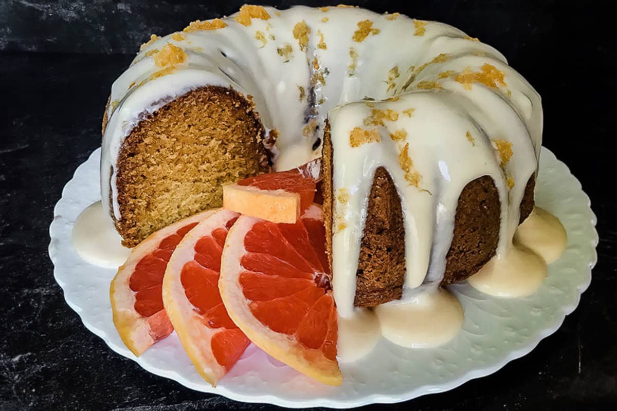 A bundt cake topped with white icing is placed on a white plate. A slice of the cake is missing, revealing its moist interior. Several slices of pink grapefruit are arranged alongside the cake as garnish. The background is dark, highlighting the dessert.