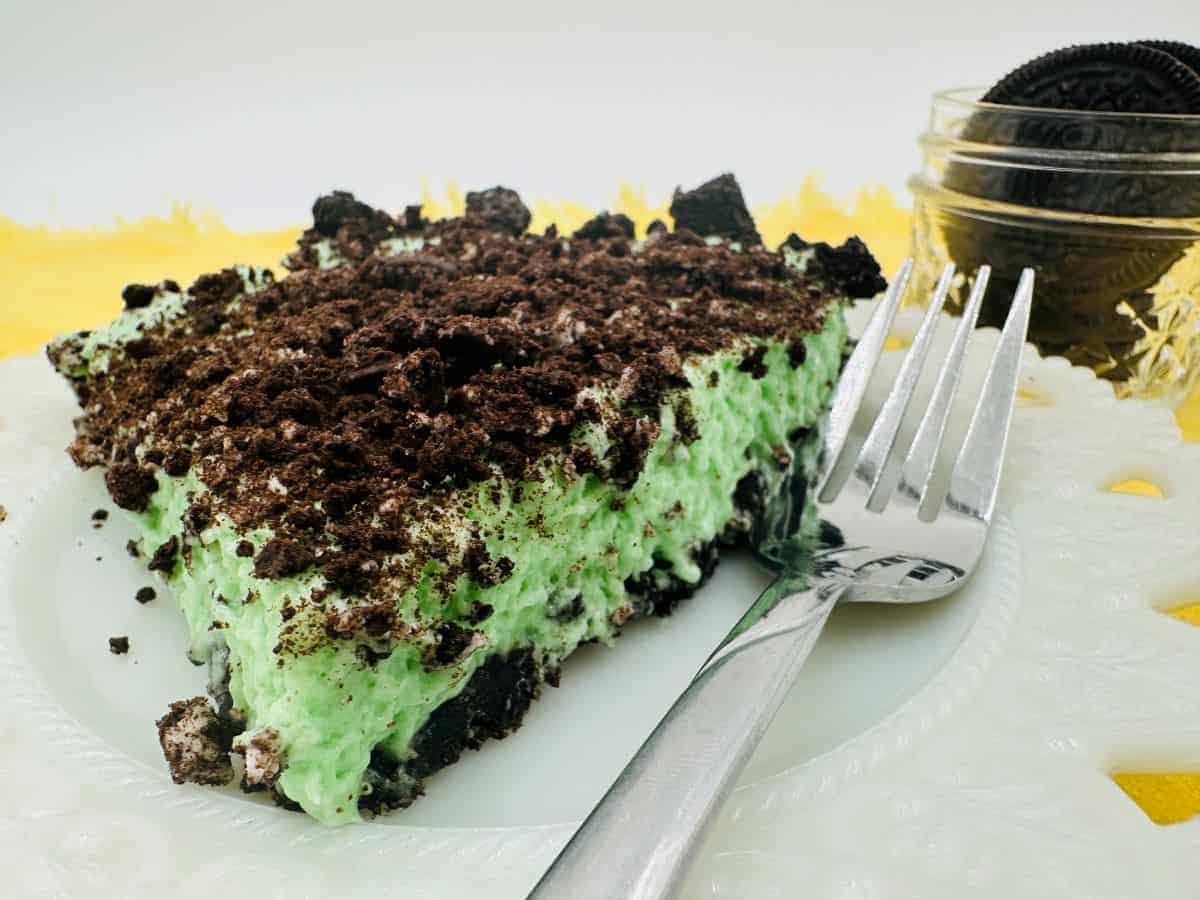 A plate with a slice of green, mint-flavored dessert topped with crushed chocolate cookies. A fork is placed beside the dessert on the plate. A jar with more chocolate cookies is in the background, with a bright yellow cloth underneath part of the plate.