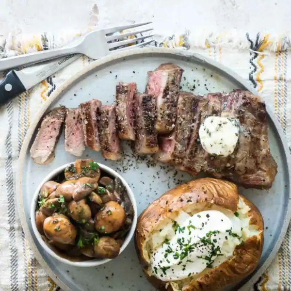A plate of sliced steak topped with butter, accompanied by a baked potato loaded with sour cream and chives, and a small bowl of seasoned mushrooms. A knife and fork rest on the edge of the plate on a checkered cloth.