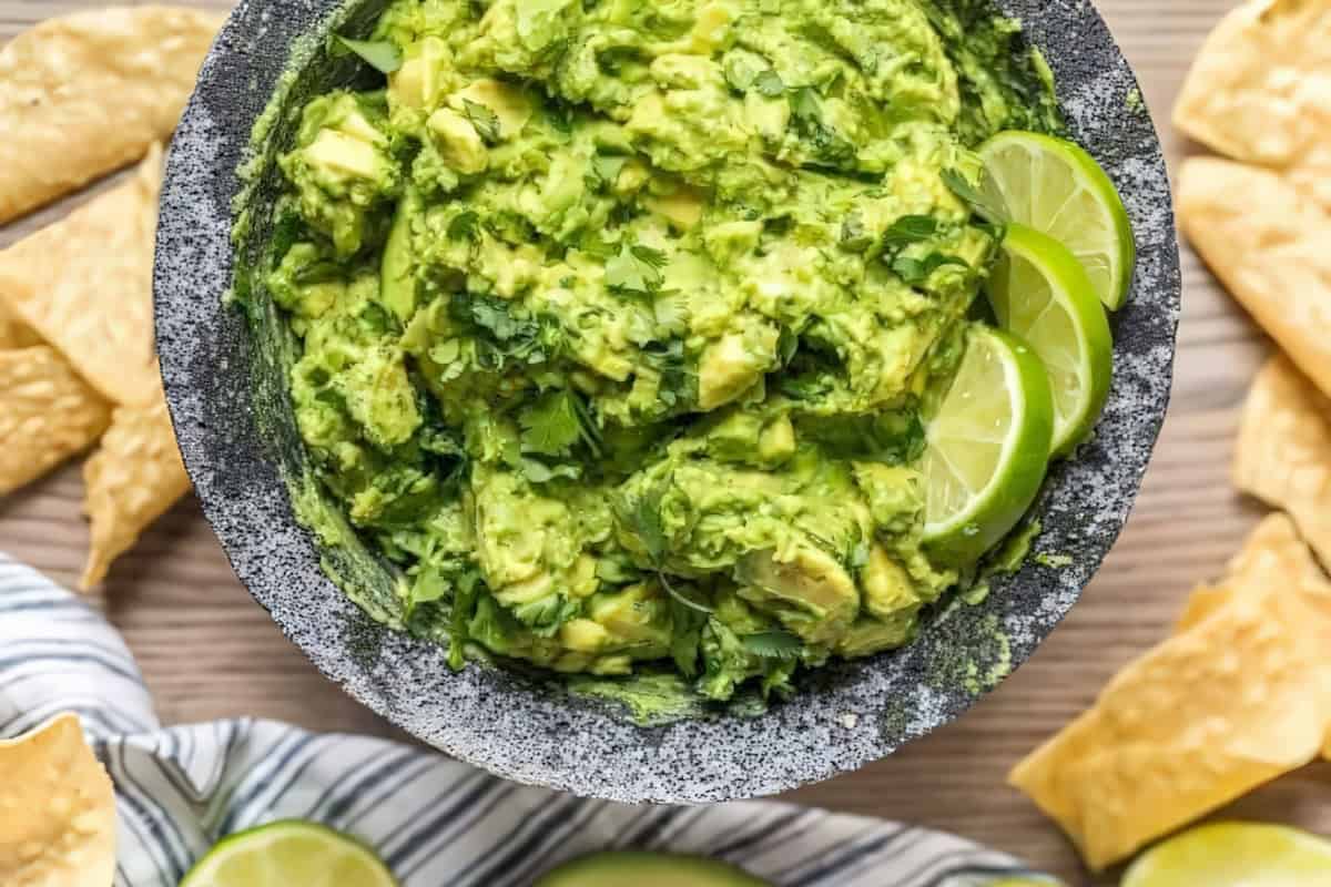 A stone bowl filled with chunky guacamole garnished with lime slices and cilantro sits on a wooden surface. Surrounding the bowl are scattered tortilla chips and part of a striped cloth.