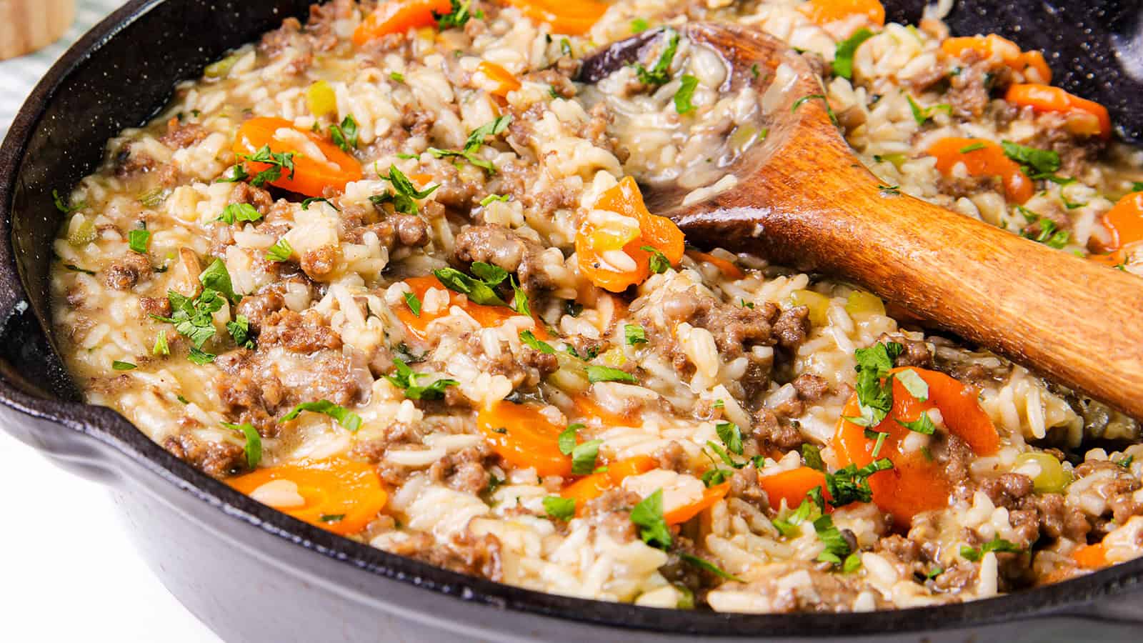 A skillet contains a mixture of rice, ground meat, diced carrots, celery, and fresh herbs. A wooden spoon rests in the dish.