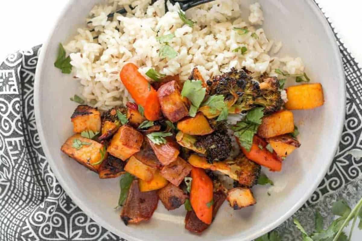 A white bowl filled with white rice, roasted vegetables including broccoli, sweet potatoes, carrots, and bell peppers, garnished with fresh cilantro. The bowl is placed on a patterned black and white napkin.