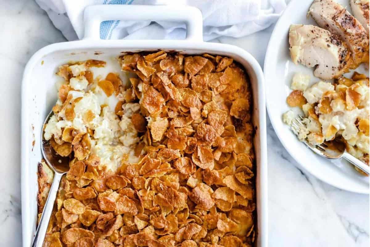 A rectangular white baking dish filled with hash brown casserole topped with crispy cornflakes, partially scooped out with a spoon. Next to it is a plate with a serving of the casserole and pieces of roasted chicken. A white cloth is in the background.
