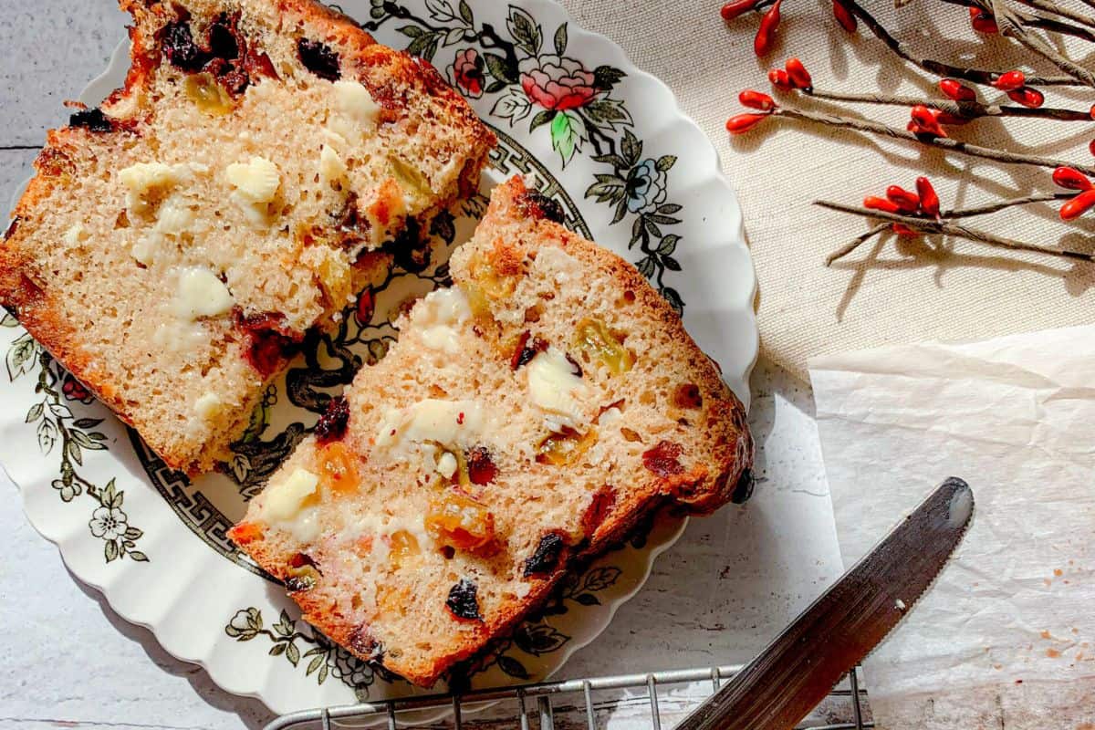 Two slices of fruit and nut bread with melted butter are on a floral plate. The plate is placed on a white surface next to a butter knife, parchment paper, and a branch with red berries. Sunlight casts a warm glow on the scene.