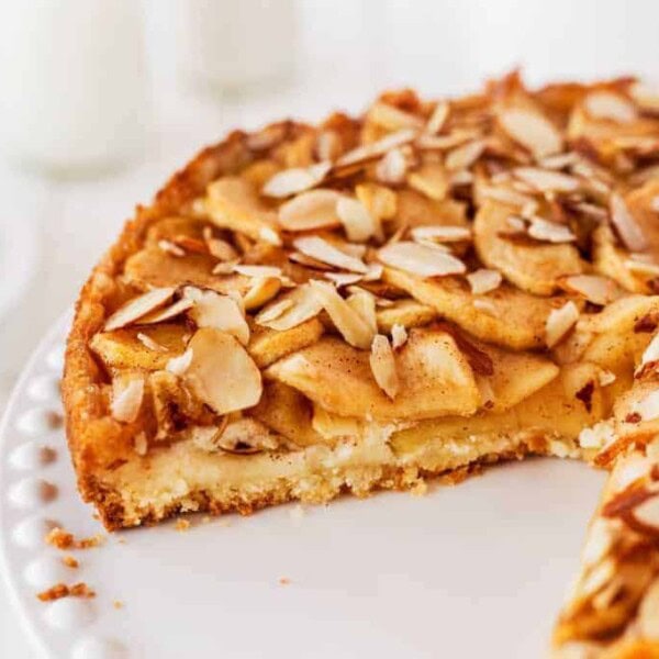 A close-up of a partially sliced apple almond cake on a white plate. The cake features a golden-brown crust, layers of sliced apples, and a topping of slivered almonds. A blurred background shows a slice of the cake on a separate plate and two glasses.