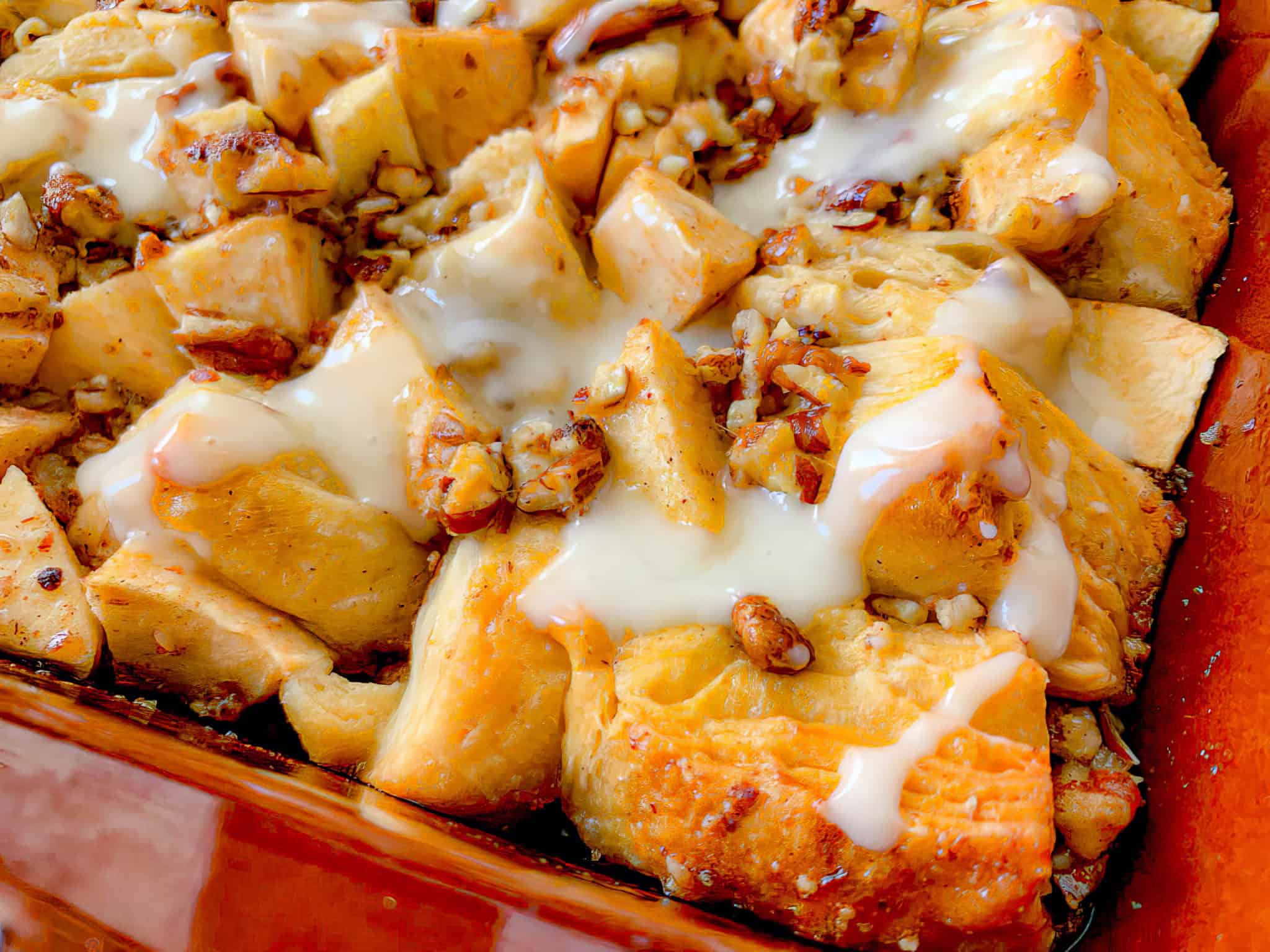 A close-up of a baked dish consisting of flaky, golden-brown chunks of bread or pastry topped with chopped nuts and drizzled with a creamy white glaze. The image shows crispy textures and hints of various ingredients mixed throughout.