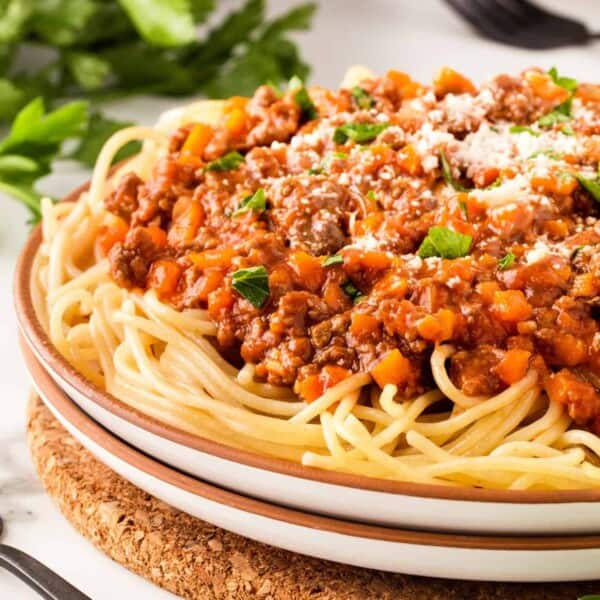 A plate of spaghetti with meat and vegetable Bolognese sauce, parsley, and grated cheese on a white table with nearby forks.