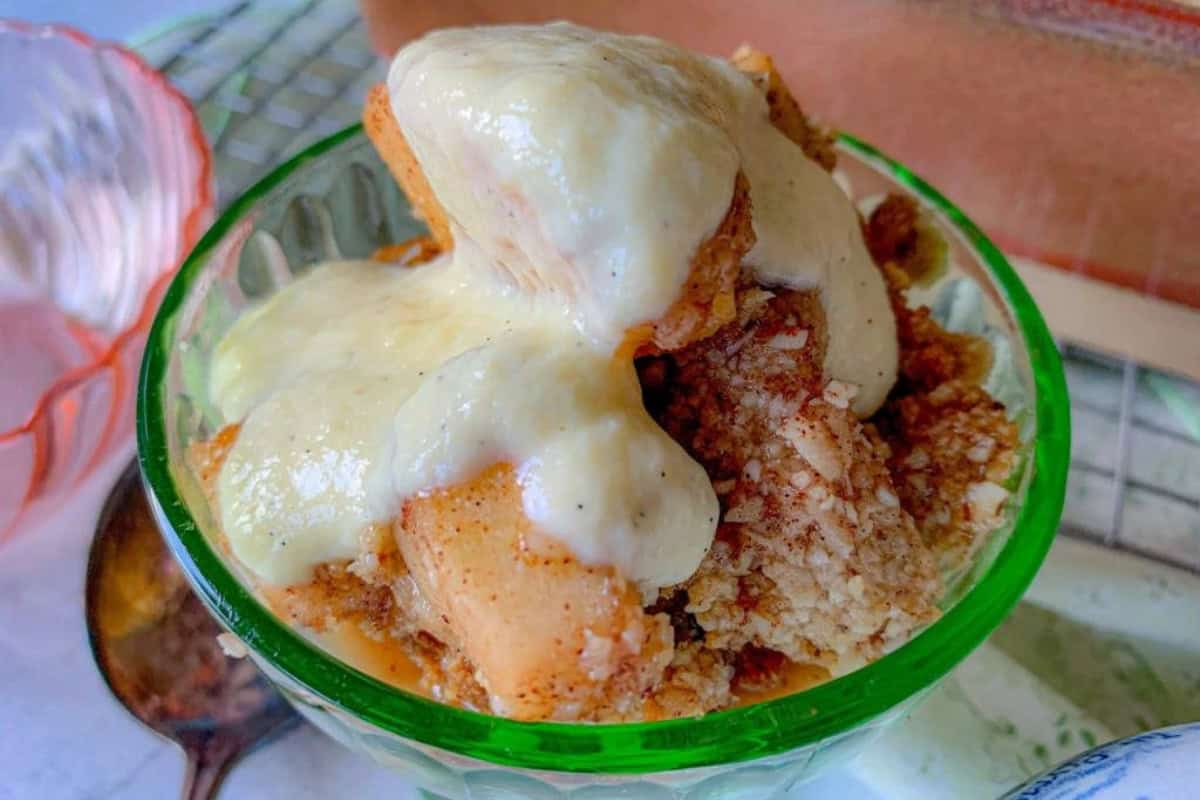 A close-up of a dessert in a green glass bowl filled with slices of apple crumble topped with a rich, creamy sauce. A spoon and an empty dish can be seen in the background.