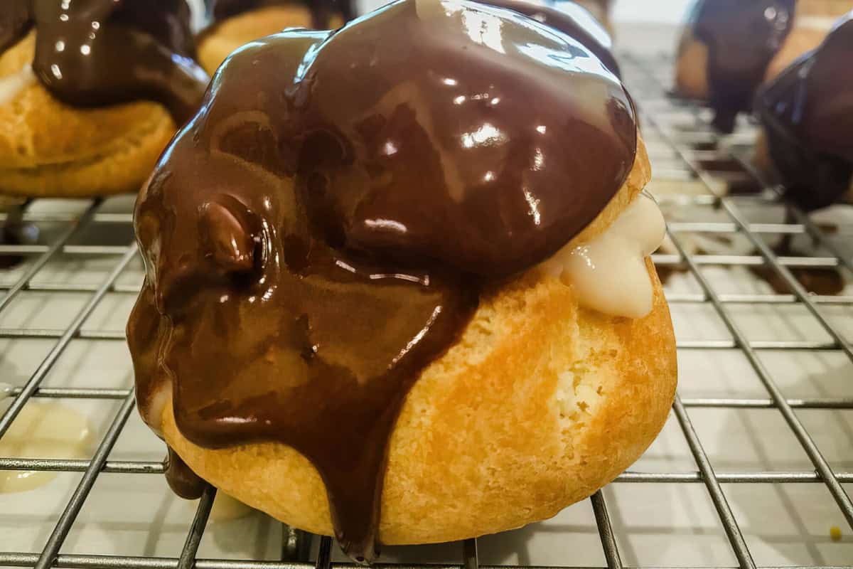 A cream puff filled with vanilla custard and topped with chocolate glaze rests on a wire cooling rack.