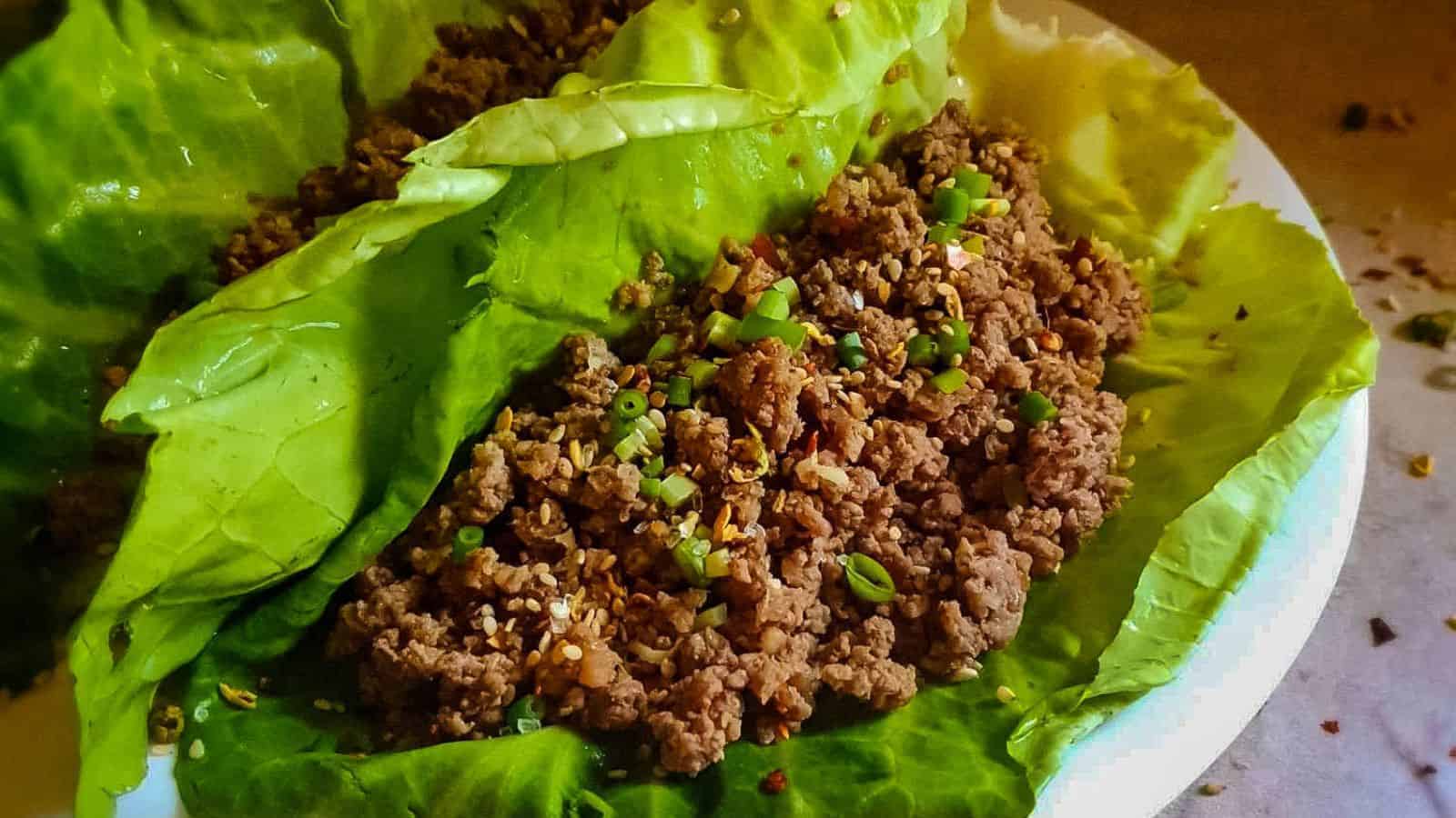 A dish of minced meat served on fresh green lettuce leaves, garnished with chopped green onions and sesame seeds, placed on a white plate.