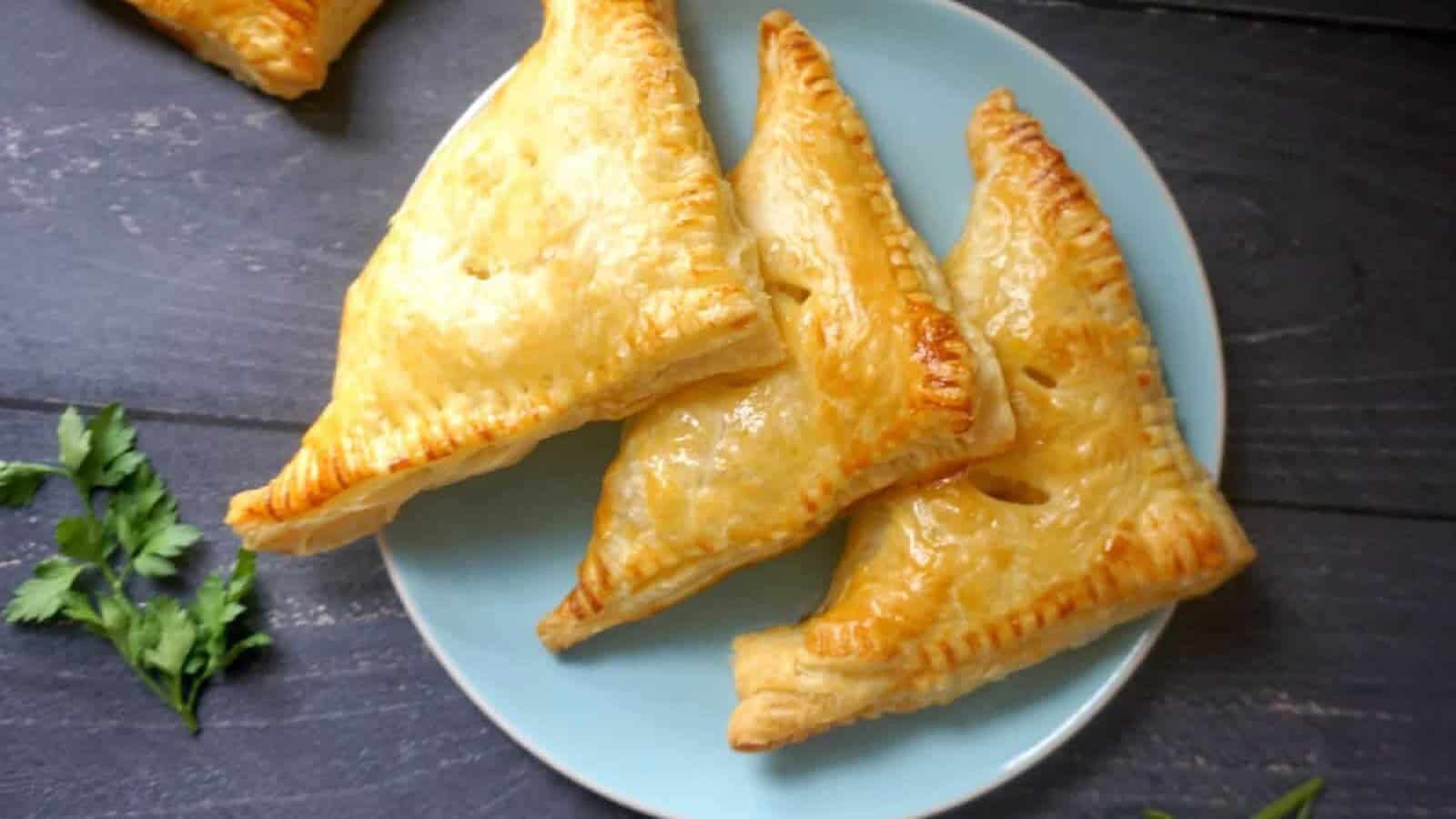 Three golden-brown puff pastry pies rest on a light blue plate, accompanied by parsley and placed on a dark wooden surface.