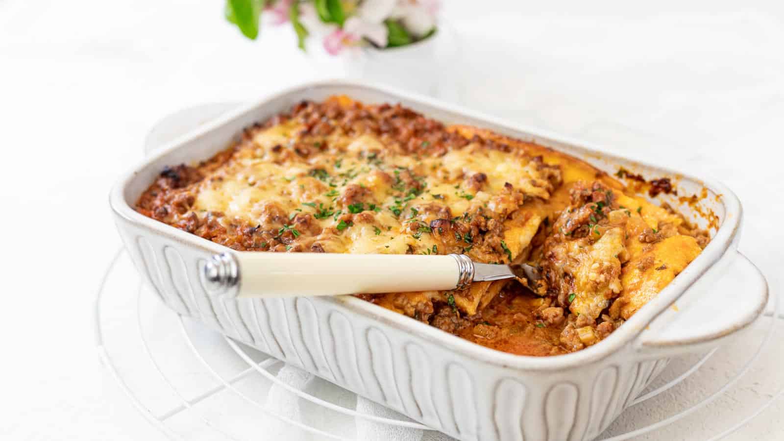 A baked lasagna in a rectangular white ceramic dish, topped with melted cheese and garnished with herbs. A slice has been cut and is being served with an ivory-handled utensil. The background features a white tablecloth and a vase with a few pink flowers.