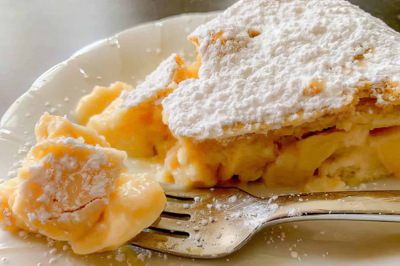 A slice of custard pie topped with powdered sugar on a white plate, with a fork holding a bite of pie.
