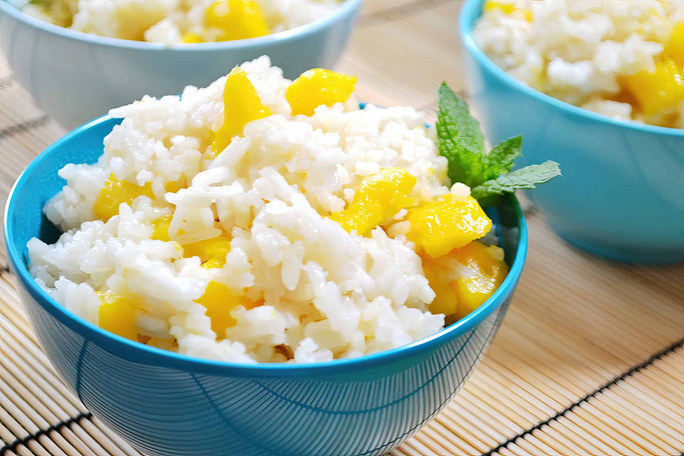 A close-up of a blue bowl filled with mango sticky rice, garnished with a sprig of mint. The dish features white sticky rice mixed with yellow mango chunks. Similar bowls are visible in the background, placed on a bamboo mat.