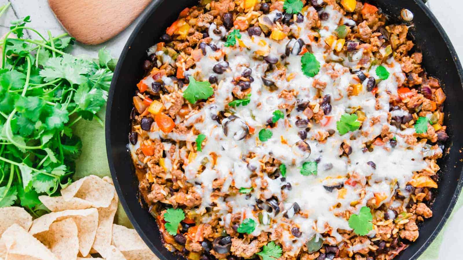 A skillet filled with a colorful mix of ground meat, black beans, corn, diced tomatoes, and melted cheese, garnished with cilantro. The skillet is placed next to fresh cilantro and tortilla chips.