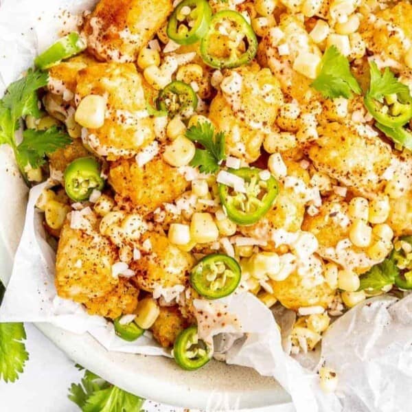 A plate of tater tots topped with corn kernels, sliced jalapeños, chopped cilantro, and Tex Mex seasoning, accompanied by a small bowl of dipping sauce.