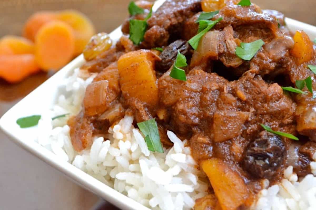 A close-up of a plate of rice topped with a rich, savory stew. The stew appears to contain chunks of tender meat, diced onions, possibly raisins, and garnished with fresh parsley. In the background, there are a few pieces of dried apricot.